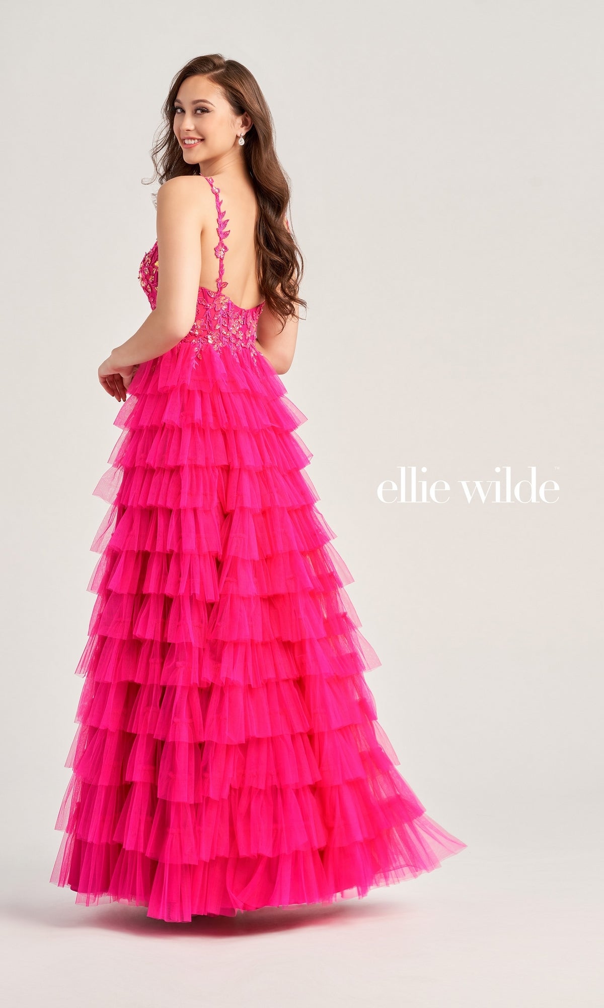 A woman smiles over her shoulder in the sleeveless, bright pink Tiered Ellie Wilde EW35059 Designer Ball Gown with a ruffled skirt. She stands against a plain light gray backdrop with